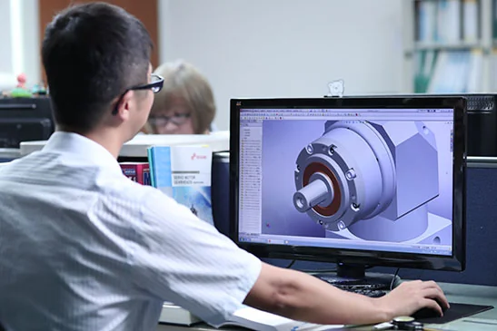 A man in a white shirt and glasses works on a computer, viewing a 3D model of a gearbox on the screen.