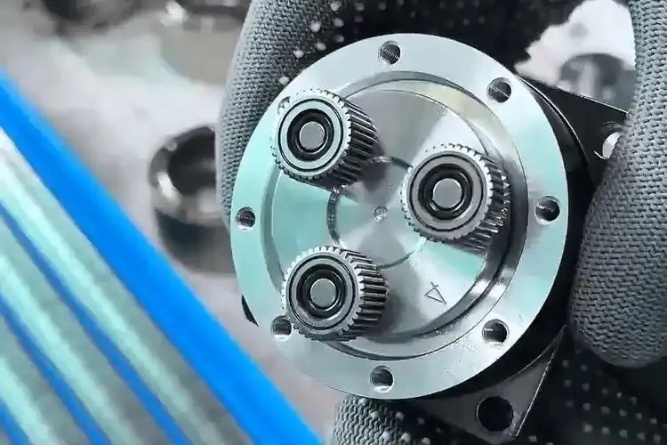 A gloved hand holds a circular metal component with three small gears and bearings on its surface, surrounded by screw holes.