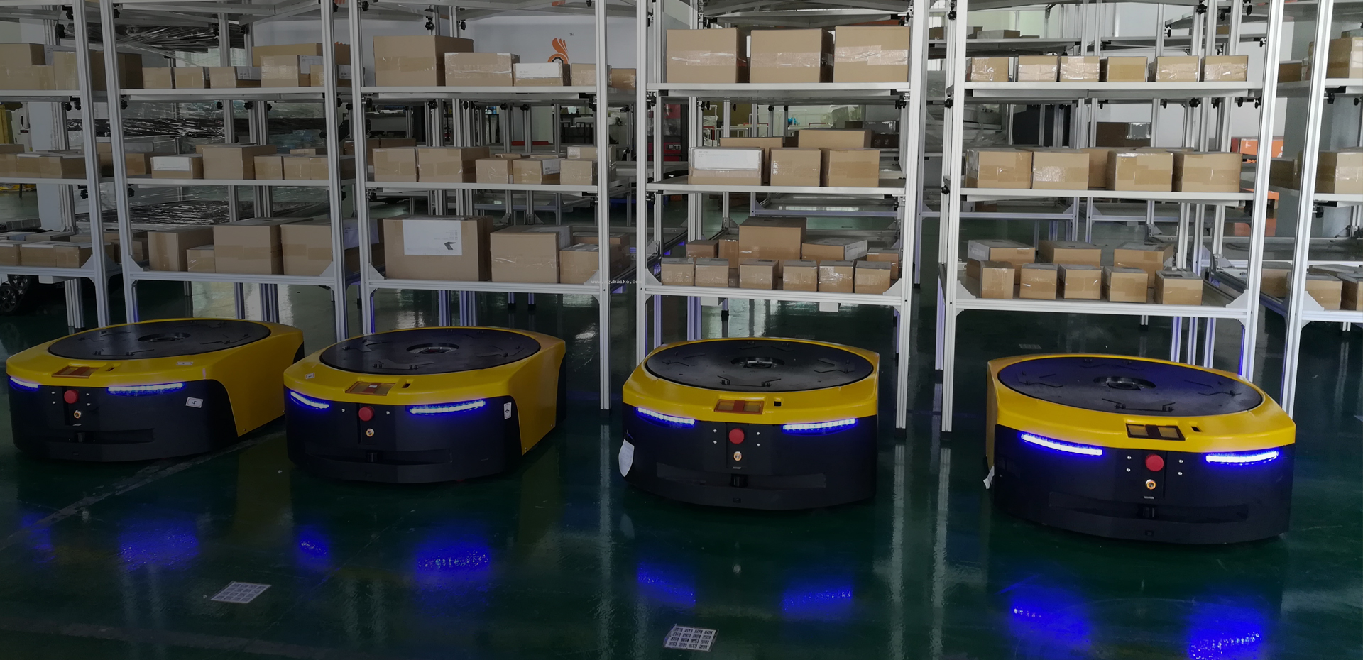 Bốn AGV tiềm ẩn với đèn xanh phát sáng xếp thành hàng trước kệ kim loại chứa đầy hộp.