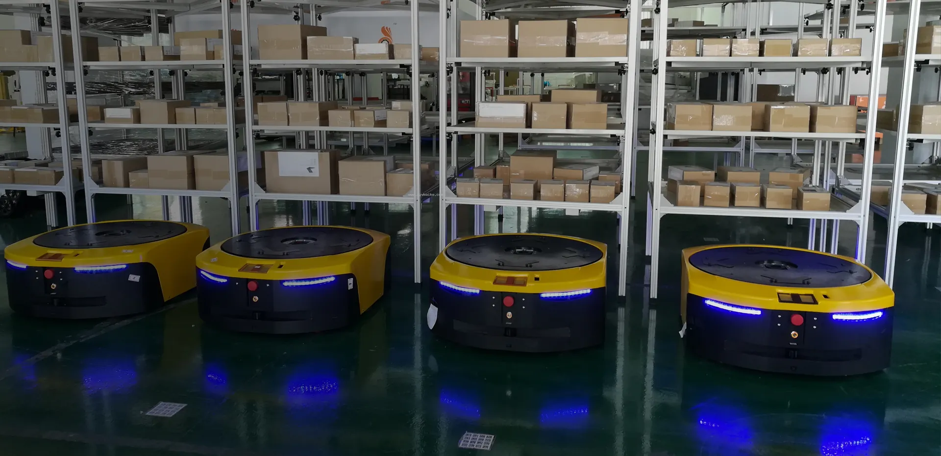 Four latent AGV with glowing blue lights in a row in front of metal shelves filled with boxes.