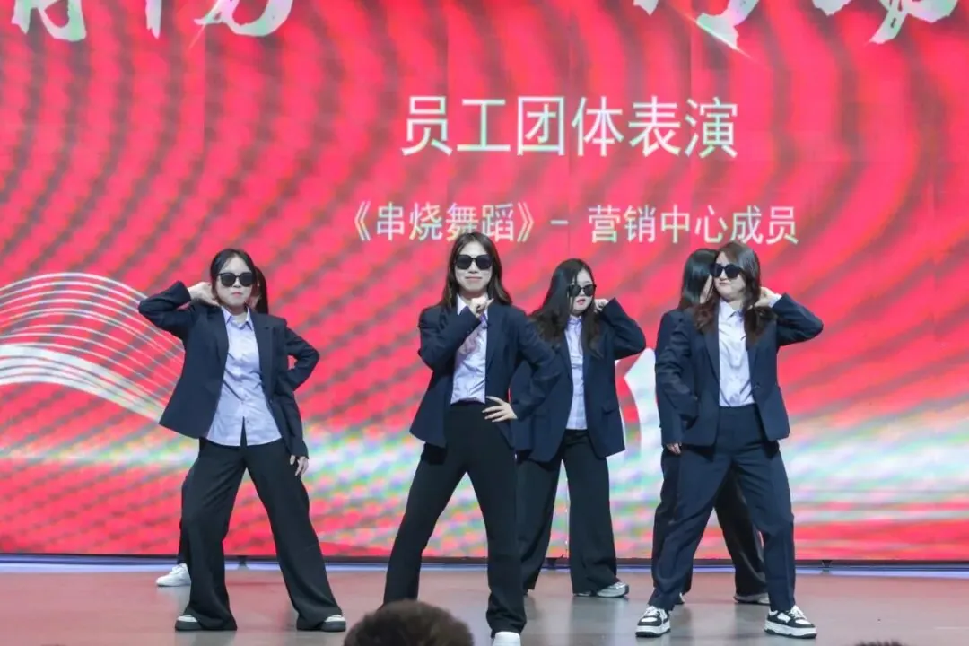 Four women in dark suits, light shirts, dark pants, and sunglasses pose on a stage against a red, wavy background.