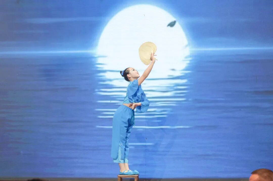 A dancer in blue attire stands on a small platform, reaching upward with a straw hat against a blue backdrop of water and a bright full moon.