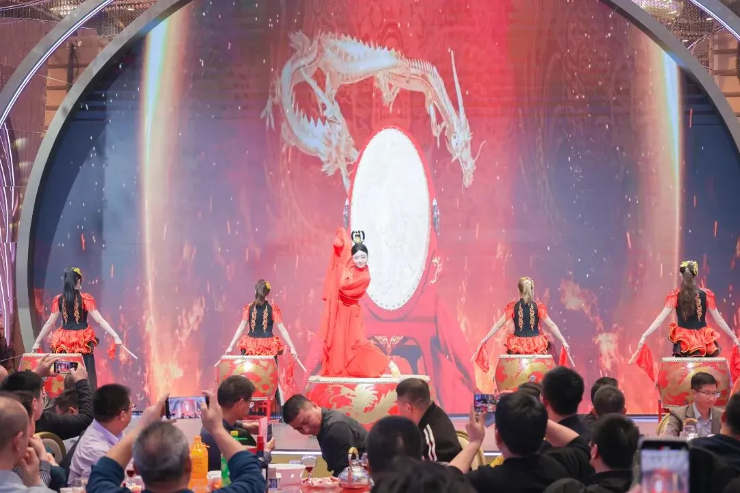 A stage performance featuring a woman in red near a large drum, with four backup performers on smaller drums, set against a backdrop of a stylized dragon and red, fiery graphics.