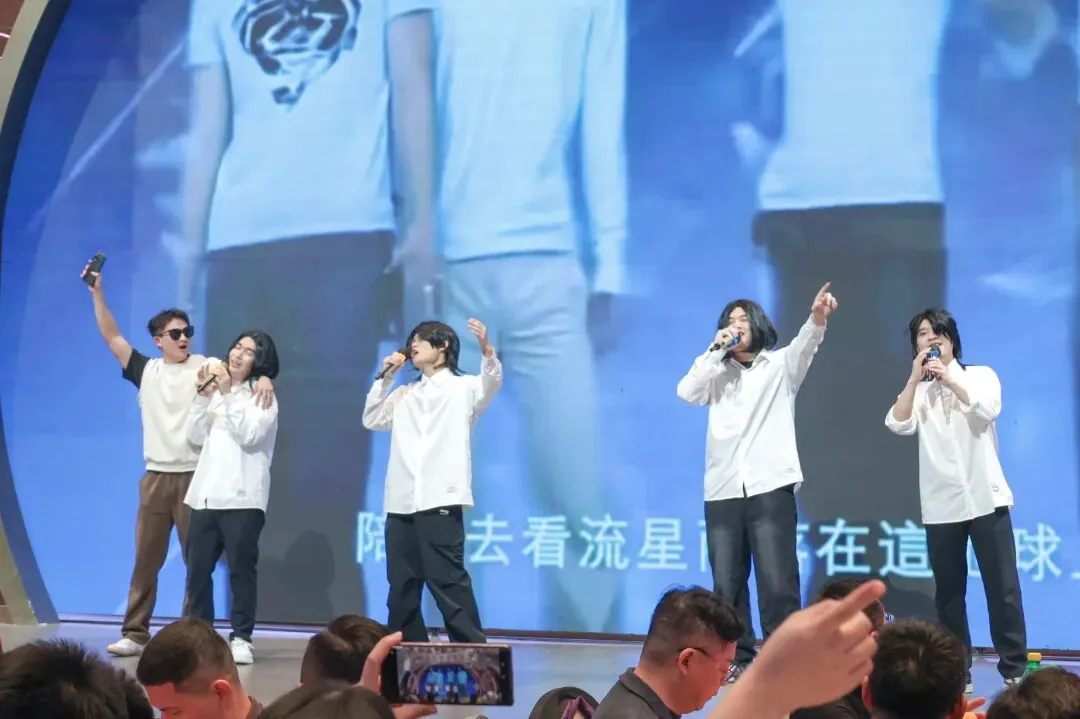 Five men on a stage, four holding microphones, perform in front of a large blue screen showing enlarged figures and some characters.